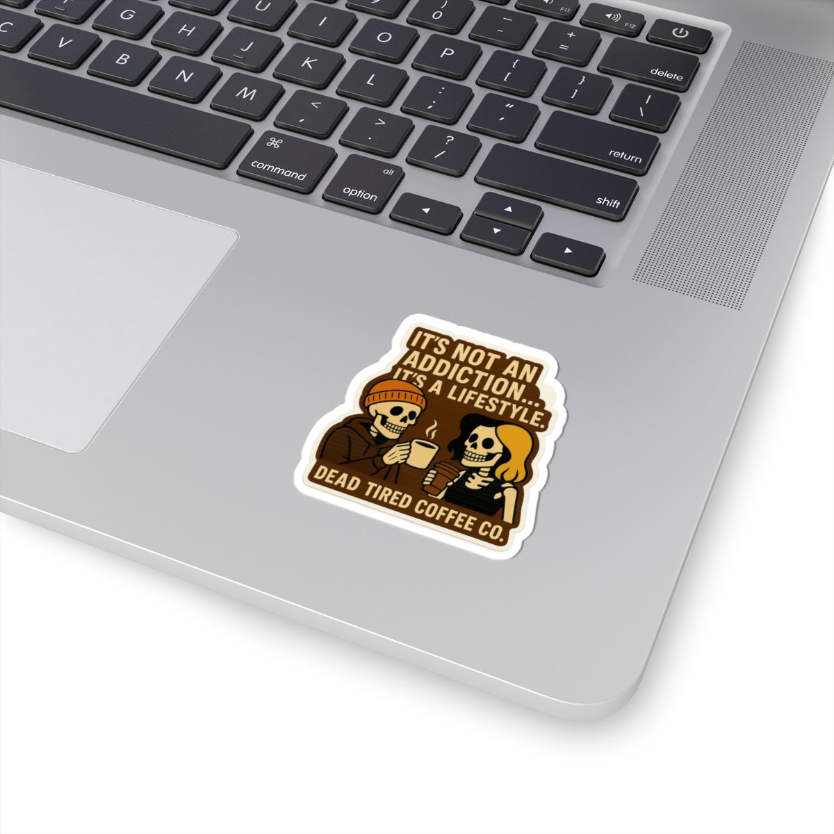 Laptop with a sticker featuring skeletons and text about coffee on a white background